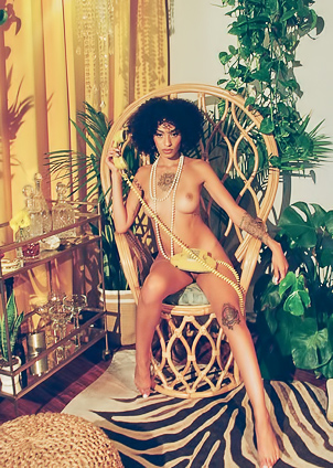Sunni in yellow lingerie poses solo on wicker chair surrounded by plants and warm light